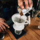 Close up of a young male barista hands pouring coffee into ceramic glass and measure it on digital scale in coffee shop. Barista wearing dark uniform. Tools and equipment for making Drip Brew coffee on wooden table.